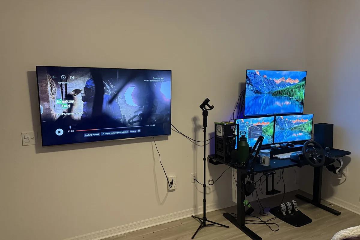 Wall-mounted TV with gaming setup featuring three monitors and racing wheel